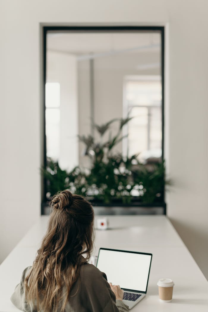 gallery-1 A woman with curly hair works on a laptop, facing a window with plants, symbolizing remote work.