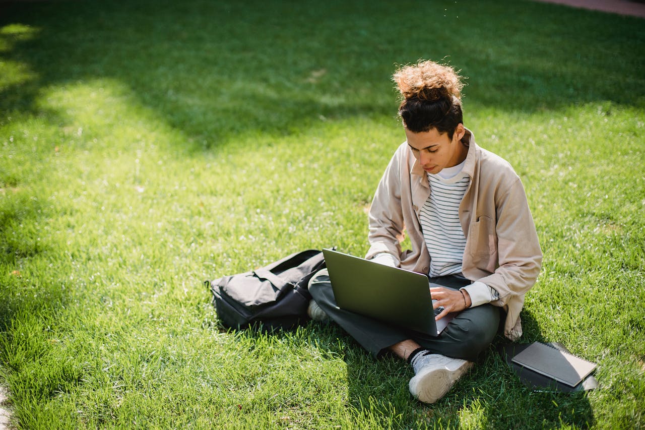 gallery-2 A young man sits on the grass studying with a laptop in a park setting.